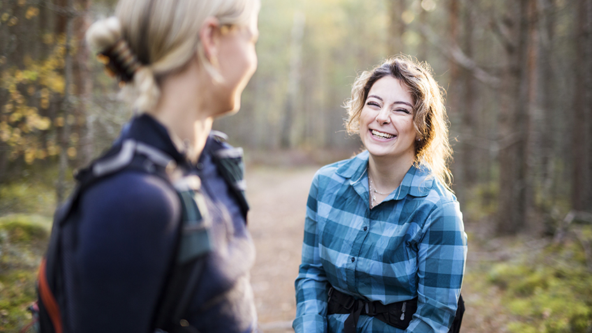Två kvinnor på vandring i skogen, har stannat upp och tittar på varandra och ler.