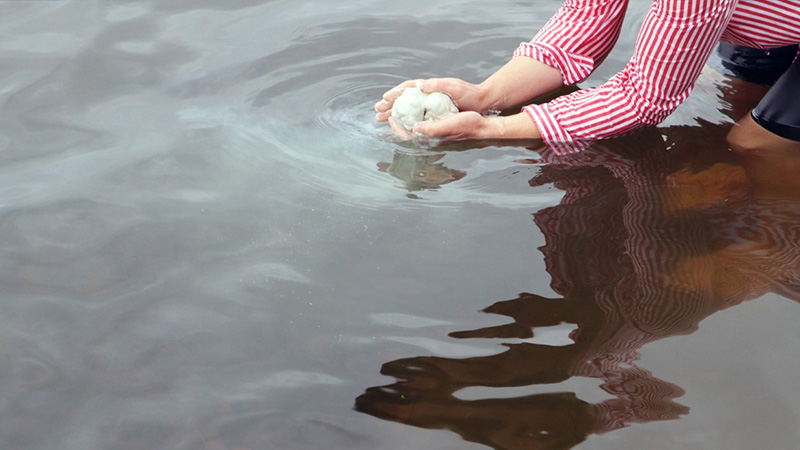 En person håller ett föremål i händerna vid vattenytan och skuggan av personen speglar sig i vattnet. 