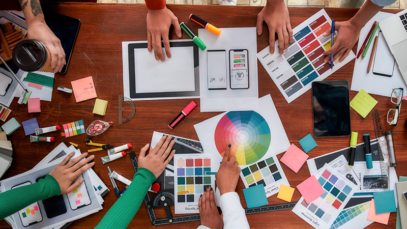 Bild ovanifrån över ett skrivbord med flera händer, färgprover, papper och pennor.