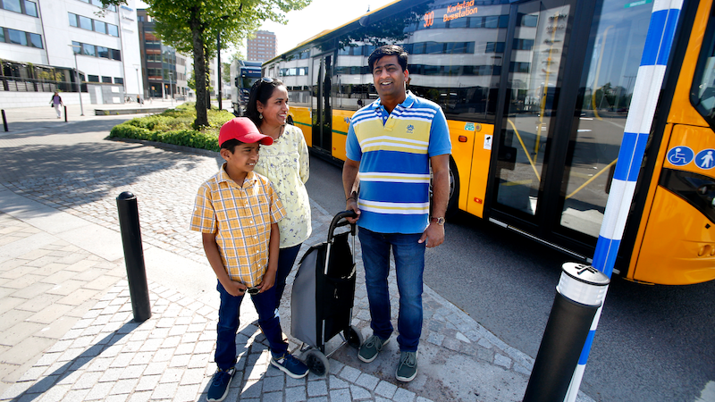 En familj, pappa, mamma och barn står vid ett övergångsställe och en gul buss åker förbi