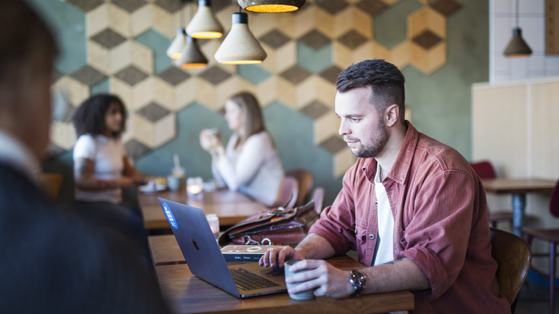 En man sitter på ett café med en dator framför sig och en kopp i handen. Vid borden runtomkring sitter personer som är i oskärpa.