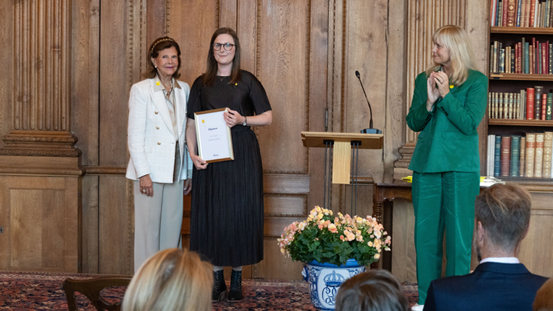 Drottning Silvia delar ut ett diplom till en medarbetare i Region Värmland. Bilden är tagen mot en mörk trävägg och i ena hörnet syns en bokhylla med böcker. En person i gröna kläder applåderar.