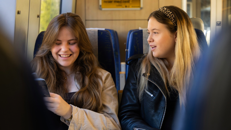 Två tjejer sitter bredvid varandra och samtalar och skrattar ombord på ett tåg.