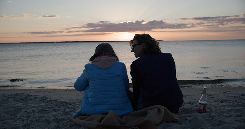 Två kvinnor på stranden. Vi ser dem bakifrån.