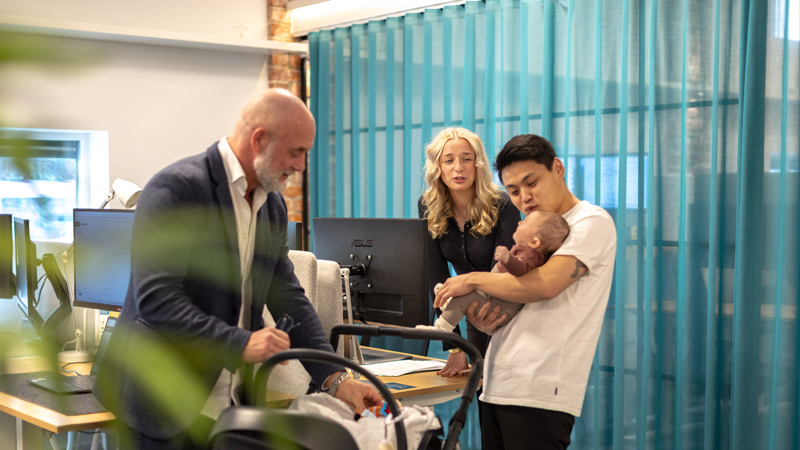 En bebis på besök i kontorsmiljö omgiven av två män och en kvinna.
