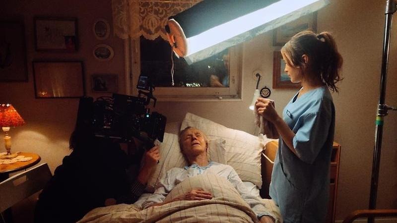 Person med filmkamera filmar äldre person i en säng och med en sköterska intill. Det är mörkt utomhus.