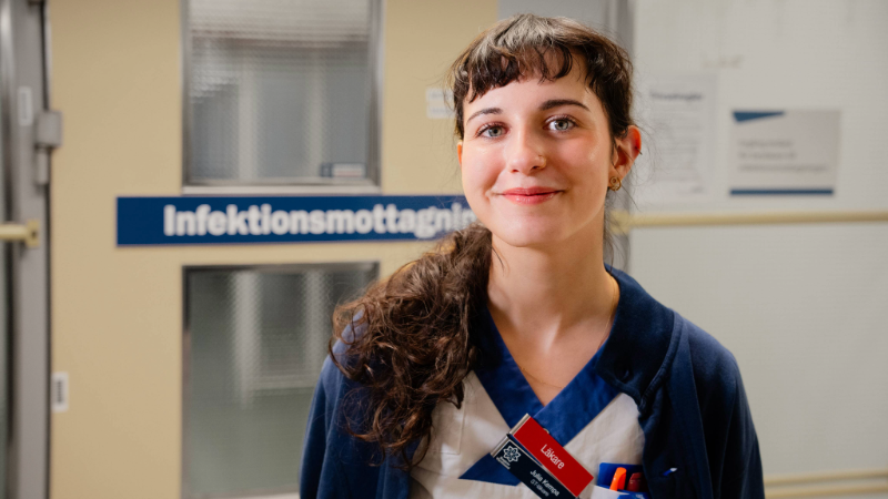 En läkare klädd i sjukvårdens vita och blåa kläder. I bakgrunden syns en dörr med texten infektionsmottagning.