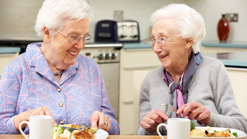 Två äldre damer äter tillsammans. De ler, tittar på varandra och pratar med varandra.