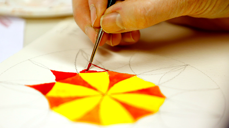 Närbild på en hand som håller i en pensel och målar akvarell i gult och rött. 