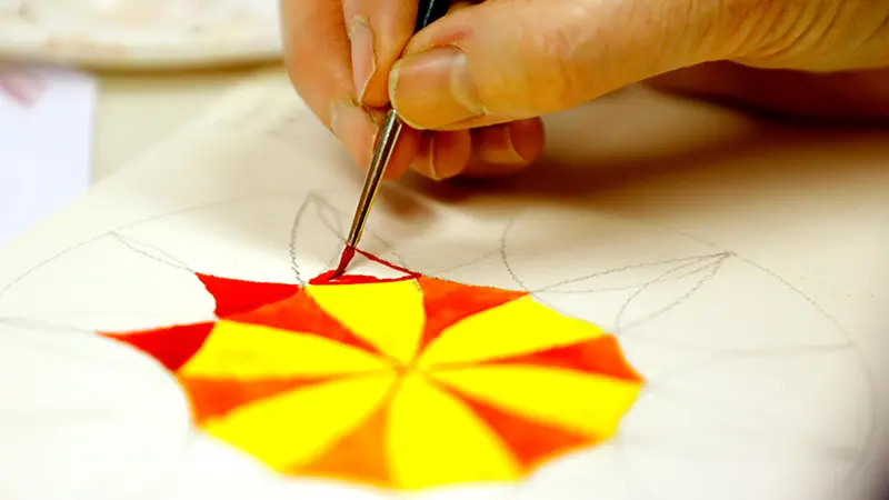 Närbild på en hand som håller i en pensel och målar akvarell i gult och rött.