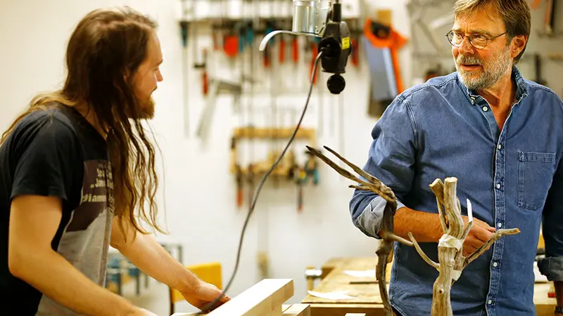 Två personer står i en verkstad med träfigurer framför sig.