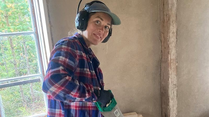 En person med hörselskydd står och tittar i i kameran med en såg i handen. 