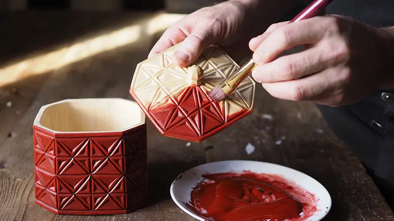 Närbild på händer som målar en snidad träask med röd färg.