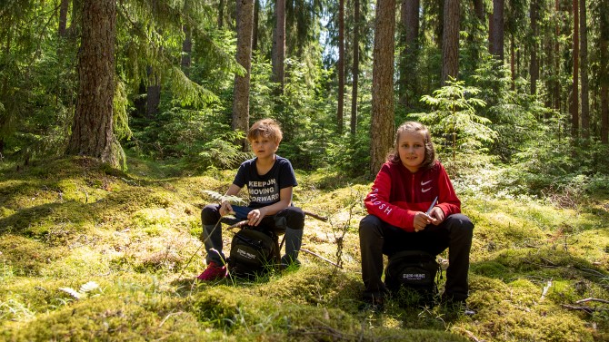 Två barn sitter på varsin ryggsäck i en solig skog.