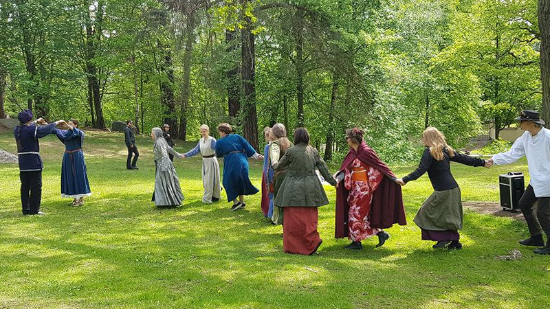 Personer i medeltida kläder som dansar hand i hand på en gräsmatta vid träd. 