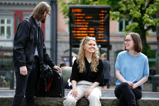 Tre ungdomar sitter och väntar på buss på Stora torget i Karlstad. I bakgrunden syns avgångstiderna för stadsbussarna i Karlstad.