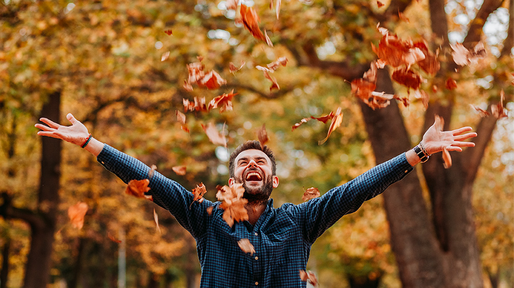 Bilden visar en glad man som kastar upp höstfärgade löv i luften.