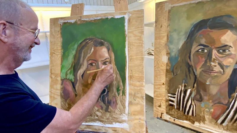 En person målar på ett av två porträtt som är uppsatta på stafflin. 