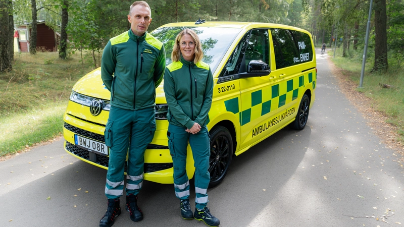 Två personer i gröna och gula kläder framför en parkerad ambulans.