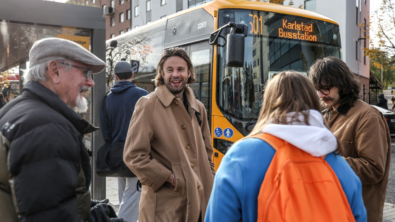 Fyra resenärer pratar glatt med varandra vid en hållplats. I bakgrunden syns en gul buss.