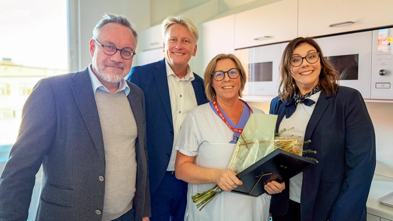 Fyra personer. En av dem har hälso- och sjukvårdens kläder och håller i en bukett och ett diplom.