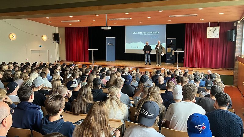 En fullsatt aula med gymnasieelever som lyssnar på två talare som står på scen.