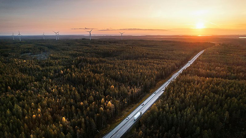 En stor väg går genom ett skogslandskap. På vägen går går både långtradare och personbilar. Till vänster i horisonten syns flera vindkraftverk och till höger går solen upp eller ner.