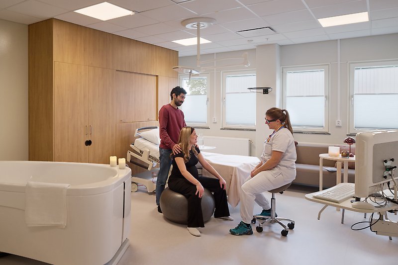 Kvinna sitter på en medicinboll, bakom stå en man, framför sitter en vårdpersonal.