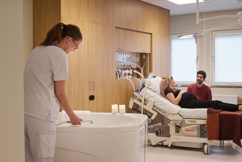Vårpersonal fyller vatten i badkar, på en säng ligger en gravid kvinna och en man sitter bredvid.